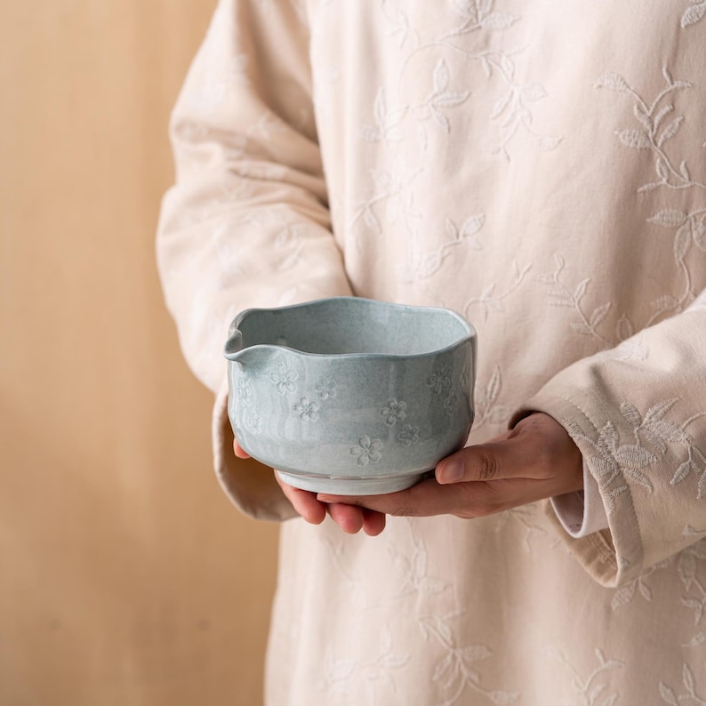 Puede incluir: Un cuenco vertedor de cer&aacute;mica azul claro con un acabado moteado y patrones florales en relieve en el exterior. El cuenco presenta un peque&ntilde;o pico vertedor en un lado y un borde redondeado ligeramente irregular, con una textura esmaltada.