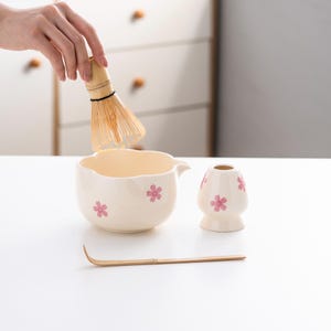 May include: A white ceramic matcha bowl with a pink floral design, a bamboo matcha whisk, a white ceramic matcha scoop, and a bamboo matcha whisk holder.