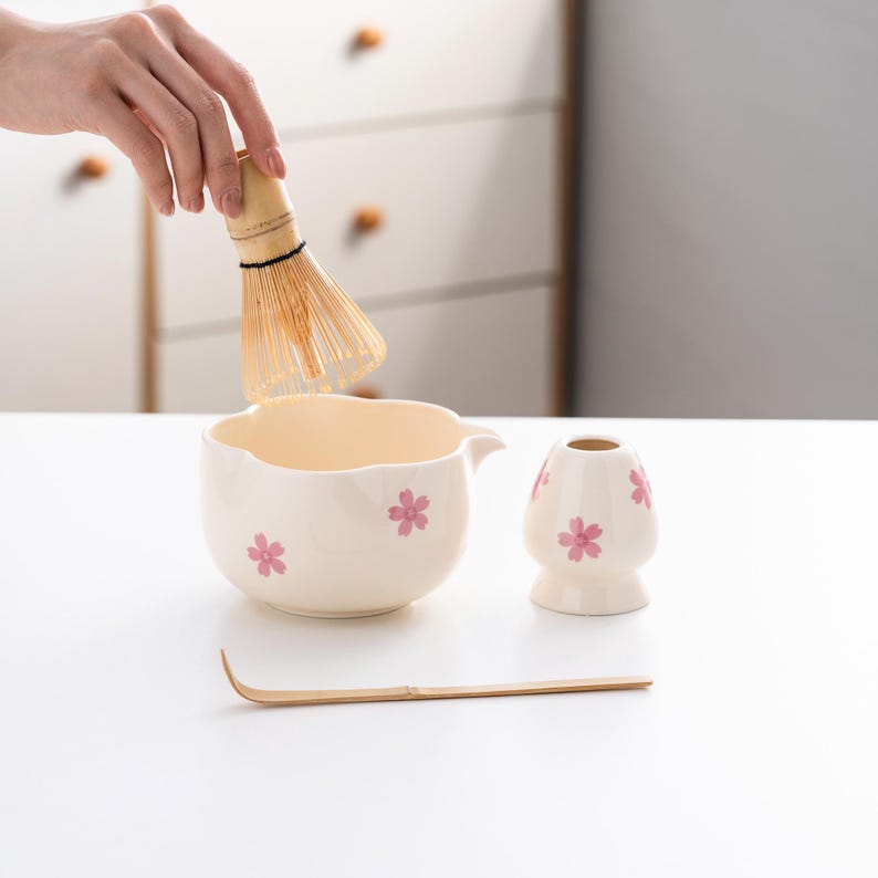 May include: A white ceramic matcha bowl with a pink floral design, a bamboo matcha whisk, a white ceramic matcha scoop, and a bamboo matcha whisk holder.