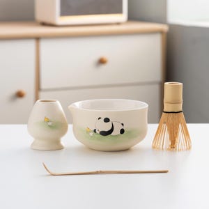 May include: A white ceramic matcha bowl with a panda bear design, a matching white ceramic whisk holder, a bamboo whisk, and a bamboo matcha scoop. The bowl has a green and white background with a black and white panda bear lying down. The whisk holder has a yellow bird design. The whisk is made of natural bamboo.