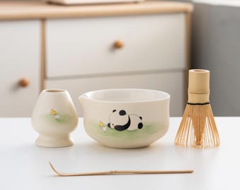 May include: A white ceramic matcha bowl with a panda bear design, a matching white ceramic whisk holder, a bamboo whisk, and a bamboo matcha scoop. The bowl has a green and white background with a black and white panda bear lying down. The whisk holder has a yellow bird design. The whisk is made of natural bamboo.