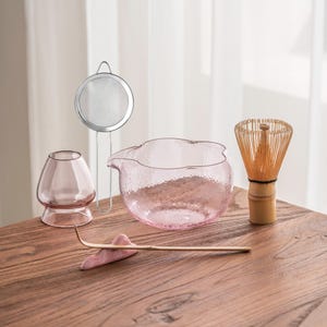 May include: A matcha tea set featuring a pink glass bowl, a bamboo whisk, a metal strainer, a pink glass cup, and a bamboo scoop. The set is arranged on a wooden surface, perfect for tea preparation.