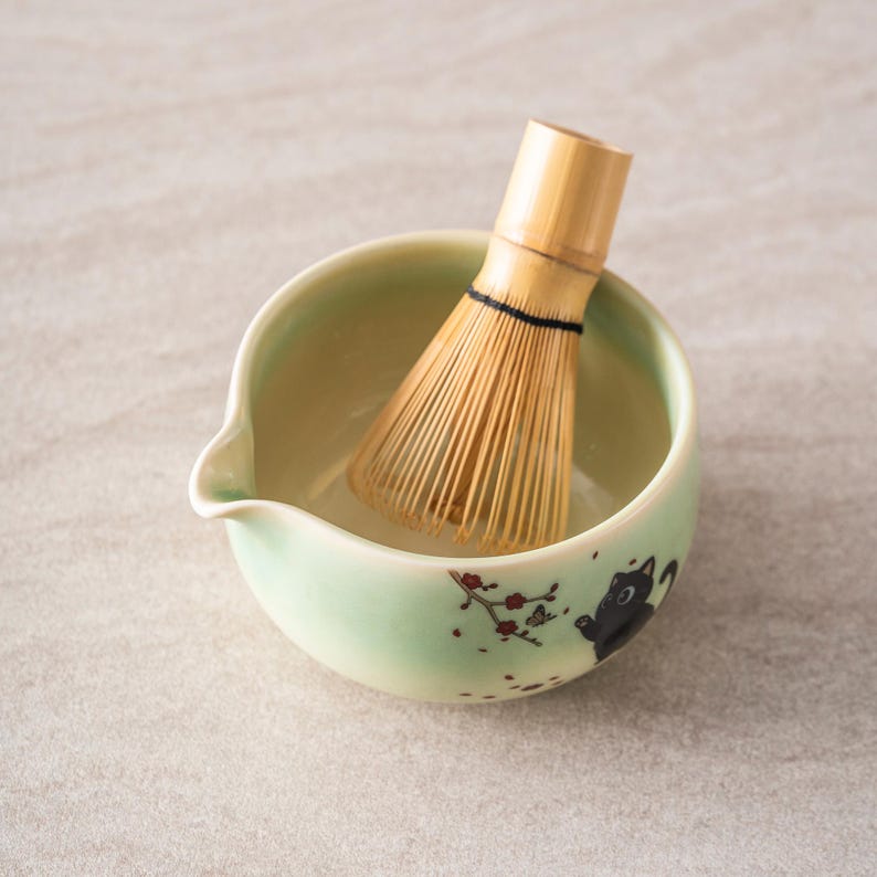 May include: A light green ceramic matcha bowl with a black cat illustration and a bamboo whisk. The bowl has a spout and a floral design. The whisk is made of thin bamboo strips.