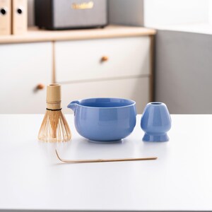 May include: A blue ceramic matcha bowl, a bamboo whisk, and a blue ceramic matcha cup on a white surface.