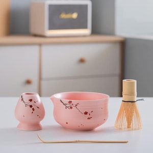 Op de afbeelding: Een roze keramische matcha-kom met een kersenbloesemontwerp, een bijpassende roze keramische gardehouder, een bamboe garde en een bamboe matcha-schep.