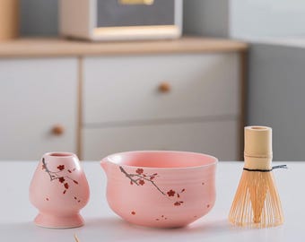 May include: A pink ceramic matcha bowl with a cherry blossom design, a matching pink ceramic whisk holder, a bamboo whisk, and a bamboo matcha scoop.