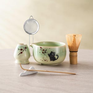 May include: A matcha tea set. It includes a light green ceramic bowl with a black cat illustration, a bamboo whisk, a ceramic tea caddy, a stainless steel strainer, and a bamboo whisk holder. The set has a floral design.