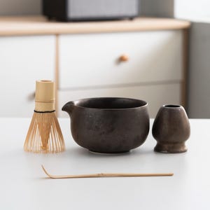 May include: A matcha tea set on a white surface. The set includes a bamboo whisk, a dark brown ceramic bowl with a spout, a small brown ceramic cup, and a bamboo scoop. A black speaker sits in the background.