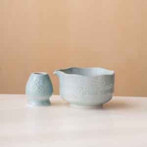 Puede incluir: Dos elementos de cer&aacute;mica azul claro para la ceremonia del t&eacute;. Un peque&ntilde;o recipiente redondeado con base acampanada y un cuenco m&aacute;s grande con borde festoneado y pico. Ambas piezas presentan un delicado estampado floral sobre un fondo neutro.