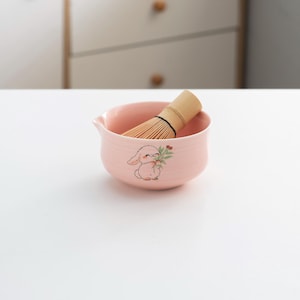 以下が含まれることがあります： かわいいウサギのデザインが施されたピンクのセラミック抹茶ボウルと、中に竹製の抹茶ホイスクが入っています