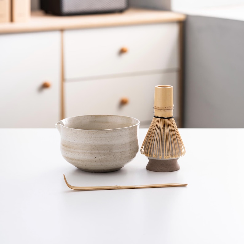 May include: A white ceramic matcha bowl with a bamboo whisk and a wooden matcha whisk holder. The bowl has a textured surface and the whisk is made of natural bamboo.