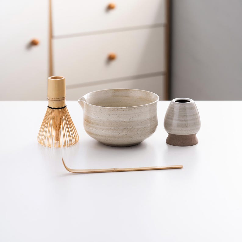May include: A white ceramic matcha bowl, a bamboo matcha whisk, a white ceramic matcha scoop, and a bamboo matcha whisk holder on a white surface.
