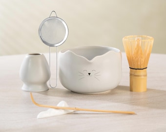 May include: A matcha tea set featuring a white ceramic bowl with a cat face design, a bamboo whisk, a tea strainer, a whisk holder, and a bamboo scoop. The set is designed for preparing matcha tea.