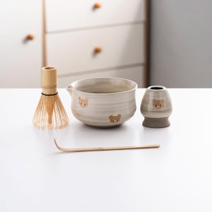 May include: A white ceramic matcha bowl with a brown bear design, a bamboo whisk, a wooden whisk holder, and a small white ceramic cup with a brown bear design. The items are on a white surface.