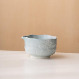 Puede incluir: Un cuenco de cer&aacute;mica azul claro con un peque&ntilde;o pico vertedor y un sutil patr&oacute;n floral blanco. El cuenco presenta una textura moteada y un borde ondulado e irregular, ofreciendo una forma distintiva para servir l&iacute;quidos o alimentos.