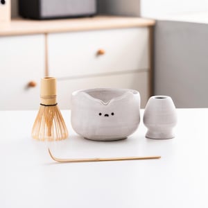 May include: A white ceramic matcha bowl with a cute cartoon face, a bamboo whisk, and a small white ceramic teacup. The set is on a white surface.