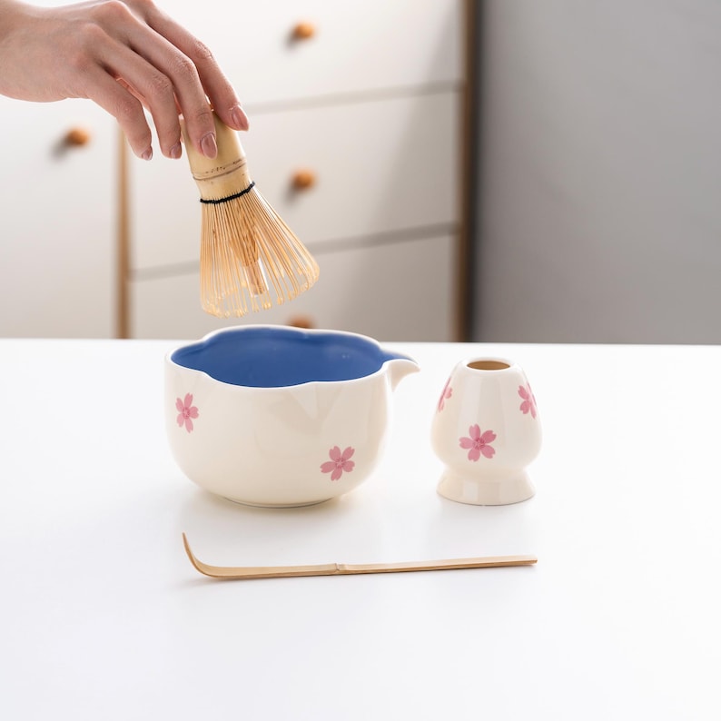 May include: A white ceramic matcha bowl with a blue interior and pink flower designs, a matching white ceramic whisk holder with pink flower designs, a bamboo matcha whisk, and a wooden matcha scoop.
