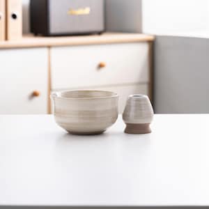 May include: A white ceramic tea bowl with a brown rim and a white ceramic tea whisk holder. The bowl and holder have a speckled glaze.