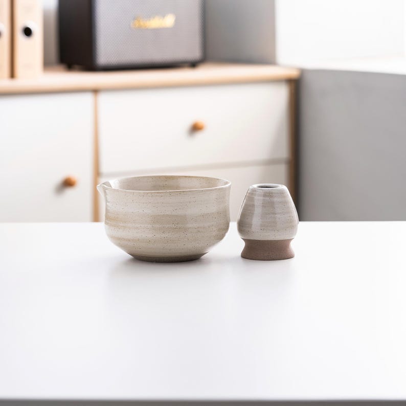 May include: A white ceramic tea bowl with a brown rim and a white ceramic tea whisk holder. The bowl and holder have a speckled glaze.