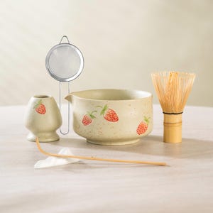 May include: A matcha tea set featuring a ceramic bowl, whisk, scoop, and a small container, all adorned with red strawberry designs. A stainless steel strainer is also included. The set is designed for preparing matcha tea.