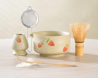 May include: A matcha tea set featuring a ceramic bowl, whisk, scoop, and a small container, all adorned with red strawberry designs. A stainless steel strainer is also included. The set is designed for preparing matcha tea.