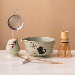 Puede incluir: Un juego de té matcha con un cuenco de cerámica con una ilustración de gato negro, un batidor de bambú, un reposacuchara de cerámica, una cuchara de bambú y un colador de acero inoxidable con un soporte de cerámica. El juego tiene un diseño floral verde y rojo.