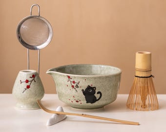 May include: A matcha tea set featuring a ceramic bowl with a black cat illustration, a bamboo whisk, a ceramic tea scoop rest, a bamboo scoop, and a stainless steel strainer with a ceramic holder. The set has a green and red floral design.