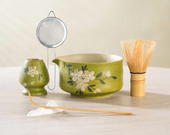 Op de afbeelding: Een matcha theeset met een groene keramische kom met bloemmotief, een bamboe klopper, een kleine keramische container, een metalen zeef en een bamboe lepel. Ontworpen voor het bereiden van matcha thee.