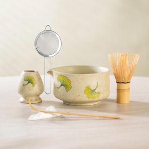 May include: A matcha tea set featuring a ceramic bowl with a spout, a bamboo whisk, a small ceramic holder, a metal sieve, and a bamboo scoop. The ceramic pieces have a speckled beige finish and are decorated with yellow ginkgo leaf designs.