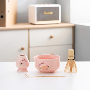 以下が含まれることがあります： 花柄のピンクのセラミック抹茶ボウルと小さなピンクのセラミック茶碗。竹製の泡立て器と竹製の抹茶スプーンも含まれています