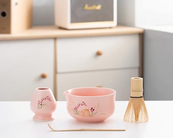 Puede incluir: Un tazón de matcha de cerámica rosa con un diseño floral y una pequeña taza de té de cerámica rosa. También se incluye un batidor de bambú y una cuchara de matcha de bambú.