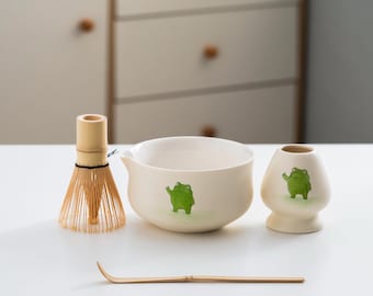 May include: A white ceramic matcha bowl with a green frog design, a bamboo whisk, a bamboo whisk holder, a white ceramic matcha cup with a green frog design, and a bamboo matcha scoop on a white surface.