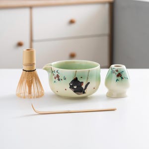 May include: A green and white ceramic matcha bowl with a black cat design, a bamboo whisk, a bamboo whisk holder, and a bamboo matcha scoop.