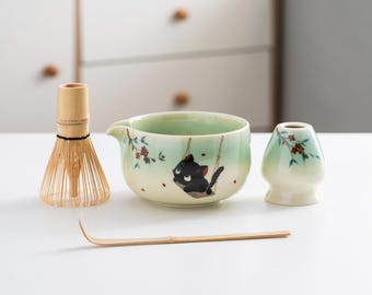 Puede incluir: Un cuenco de matcha de cerámica verde y blanco con un diseño de gato negro, un batidor de bambú, un soporte para batidor de bambú y una cuchara de matcha de bambú.