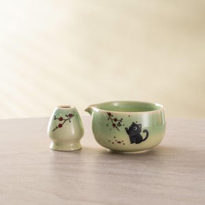 May include: A light green ceramic tea set featuring a small bowl with a spout and a whisk holder. The bowl has a black cat illustration and floral accents. The whisk holder and bowl are decorated with red and brown floral branches.