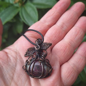Puede incluir: Un collar con colgante artesanal con un diseño envuelto en alambre. El colgante tiene una piedra preciosa de color púrpura oscuro encerrada en alambre color cobre. El collar tiene un cordón negro.