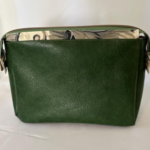 May include: A green faux leather bag with a patterned fabric top and a bronze zipper. The bag has a rectangular shape with a textured surface and bronze hardware on the sides. The patterned fabric features a mix of black, gray, and beige.