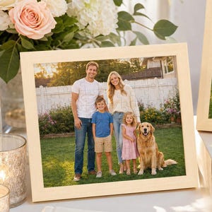 May include: Two light-colored wooden picture frames containing identical photographs of a family of four and a golden retriever standing in a grassy yard. The frames are displayed near a white guest book and a gift box tied with a satin ribbon.