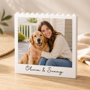 Puede incluir: Un marco de fotos rectangular blanco construido con bloques de construcción de plástico entrelazados. El marco muestra una fotografía de una persona sonriente abrazando a un golden retriever. El texto Olivia & Sunny está impreso en negro.