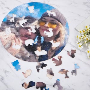 May include: A circular photo puzzle showing two people and a cat. Several animal-shaped pieces are missing from the puzzle. Additional animal-shaped puzzle pieces are scattered around. A magazine and flowers are in the background.