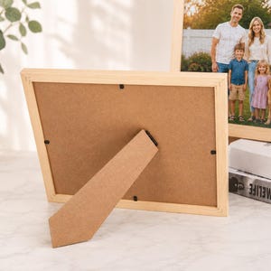 Puede incluir: Un marco de fotos rectangular de madera clara visto desde atr&aacute;s, con un respaldo de tablero de fibra marr&oacute;n y un soporte articulado. El marco contiene una fotograf&iacute;a de una familia y un perro de pie al aire libre frente a una valla blanca.