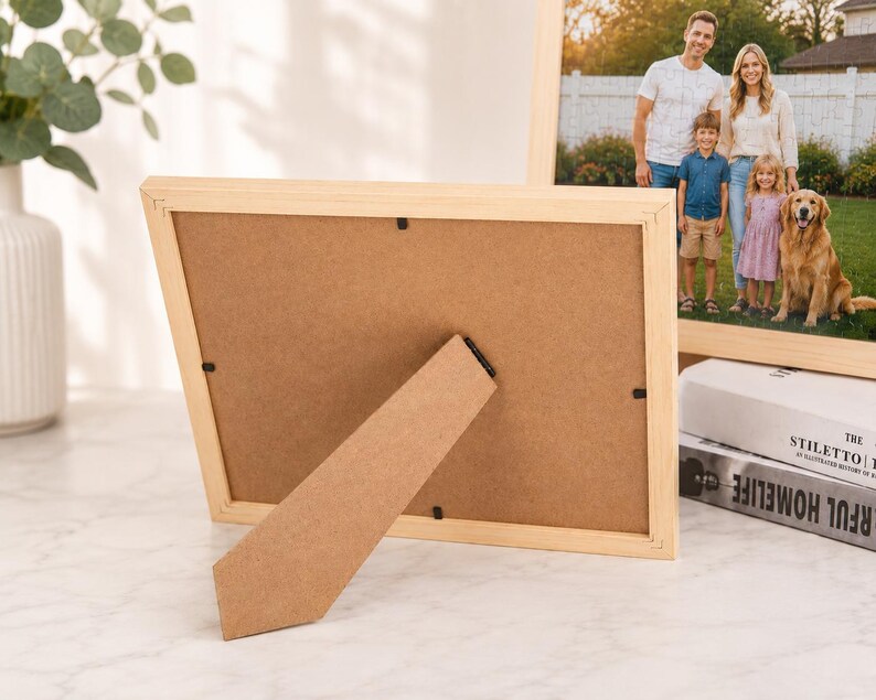 Puede incluir: Un marco de fotos rectangular de madera clara visto desde atr&aacute;s, con un respaldo de tablero de fibra marr&oacute;n y un soporte articulado. El marco contiene una fotograf&iacute;a de una familia y un perro de pie al aire libre frente a una valla blanca.