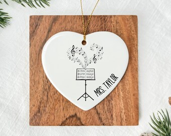 May include: A white heart-shaped ornament with musical notes and a music stand design. The ornament has a gold string for hanging and the text "MRS. TAYLOR" in black. It rests on a wooden square, with pine needles and pine cones in the background.
