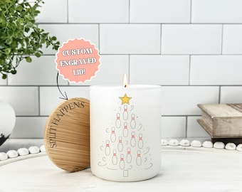May include: White candle with a bowling pin Christmas tree design. The candle has a wooden lid with the text "Split Happens". A sticker above the lid says "Custom Engraved Lid!". The candle is lit with a yellow flame.