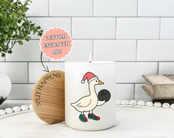 May include: A white candle with a cartoon goose wearing a Santa hat and bowling shoes, holding a bowling ball. The candle has a wooden lid with the text "Split Happens" and a pink sticker that says "Custom Engraved Lid!"