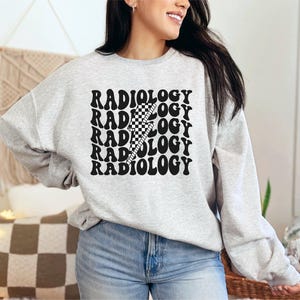 May include: A light gray sweatshirt with the word "RADIOLOGY" stacked vertically in a wavy font. A black and white checkered lightning bolt graphic is centered over the text. The sweatshirt is long-sleeved and has a crew neck.