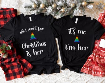Könnte beinhalten: Zwei schwarze T-Shirts mit einem Regenbogen-Weihnachtsbaum-Design. Ein Shirt sagt "alles was ich zu Weihnachten will ist sie" und das andere sagt "es bin ich, ich bin sie".