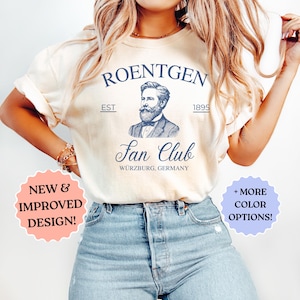 May include: Cream-colored t-shirt with a navy blue graphic. The graphic includes the word "ROENTGEN", a portrait, and the words "Fan Club" and "Würzburg, Germany". The shirt also has the text "NEW & IMPROVED DESIGN!" and "+ MORE COLOR OPTIONS!"