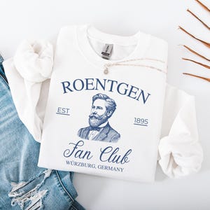 May include: A white sweatshirt with the text "ROENTGEN Fan Club Würzburg, Germany" in navy blue, along with a portrait of a man. A silver necklace with a pendant rests on the shirt, next to a pair of blue jeans.