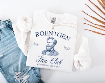 May include: A white sweatshirt with the text "ROENTGEN Fan Club Würzburg, Germany" in navy blue, along with a portrait of a man. A silver necklace with a pendant rests on the shirt, next to a pair of blue jeans.